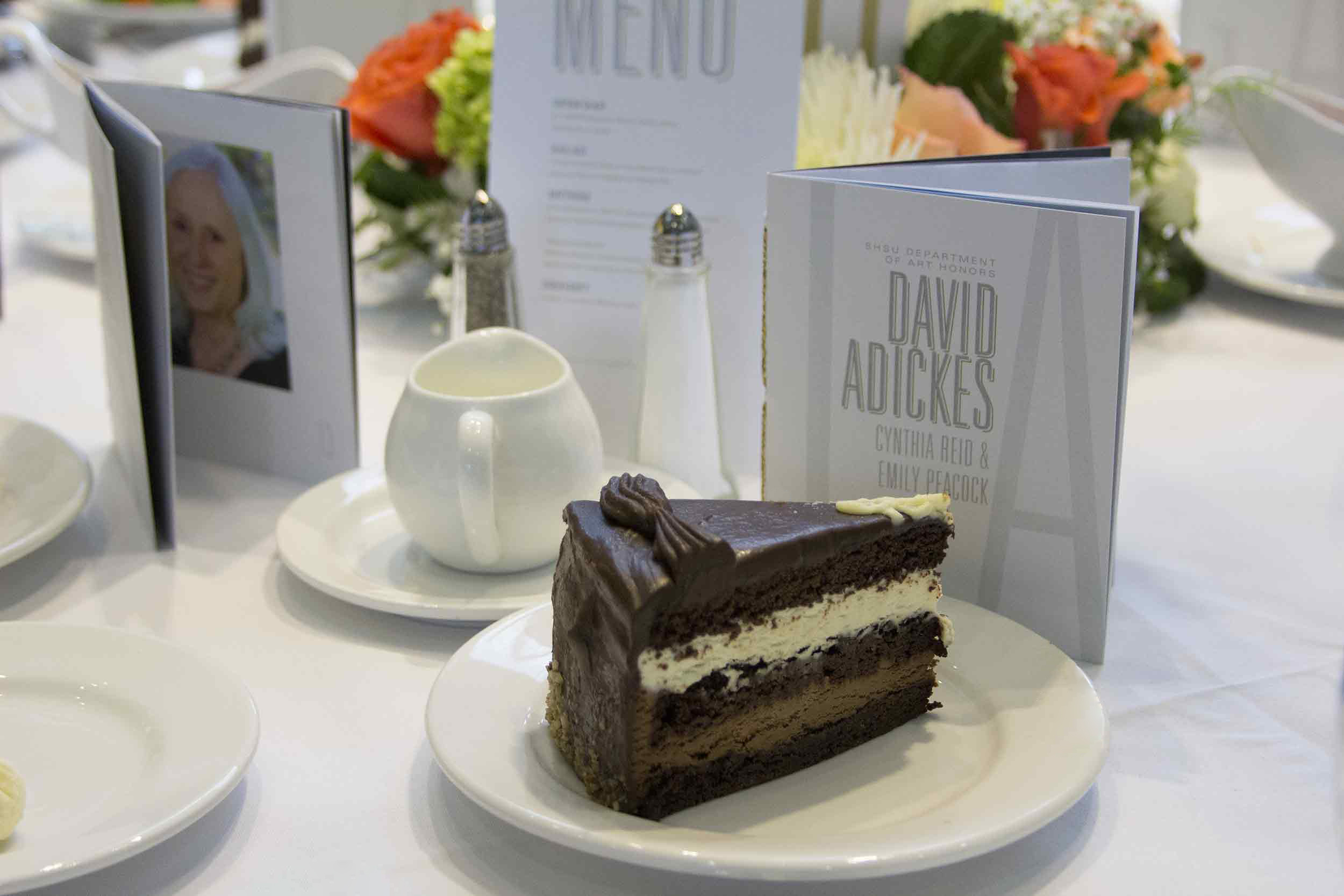 Triple layer choclate cake on a white plate with white linens with gala program and menu on a table adjacent to a vibrant floral arrangement